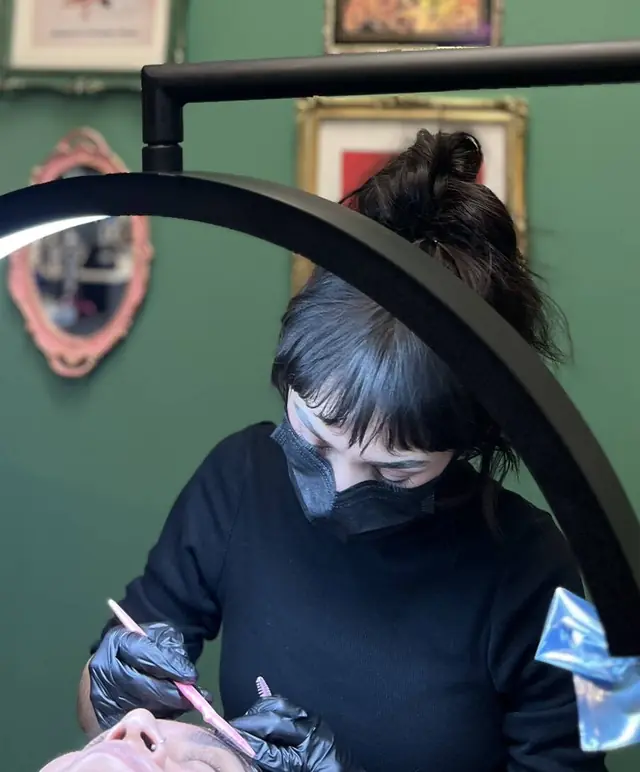 Katie, masked and gloved, working on a client's brows under an arched lamp, with gilt-framed art and a pink mirror on the green wall behind her