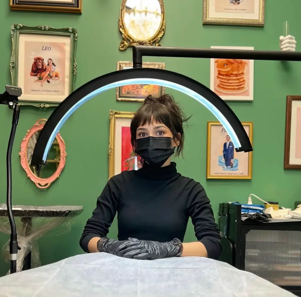 Katie McMahon in a black turtleneck, mask and gloves, seated at her treatment table under a curved lamp, with framed art and gilt mirrors on the green wall behind her